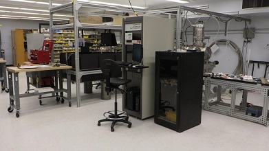 A data collection station with a computer and two racks are seen. A vacuum system is slightly visible above the smaller rack. The optical table and some benches are also visible.
